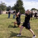 The image shows U.S. Army personnel conducting physical training exercises on a grass field. Two soldiers in the foreground wearing black Army PT (Physical Training) uniforms are performing lunges or stretching exercises with hands behind their heads. Several observers, including civilians and other soldiers, watch from the background near shipping containers and a chain-link fence.