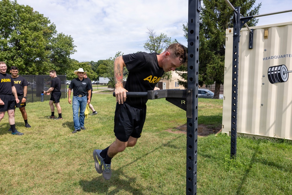 A U.S. Army soldier in black PT (physical training) uniform is performing a dip exercise on an outdoor power rack on a grassy field. Several other soldiers and a civilian in a cowboy hat observe in the background. The setting appears to be a military installation fitness area, with a shipping container labeled 'HEADQUARTERS' visible on the right.