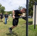 A U.S. Army soldier in black PT (physical training) uniform is performing a dip exercise on an outdoor power rack on a grassy field. Several other soldiers and a civilian in a cowboy hat observe in the background. The setting appears to be a military installation fitness area, with a shipping container labeled 'HEADQUARTERS' visible on the right.