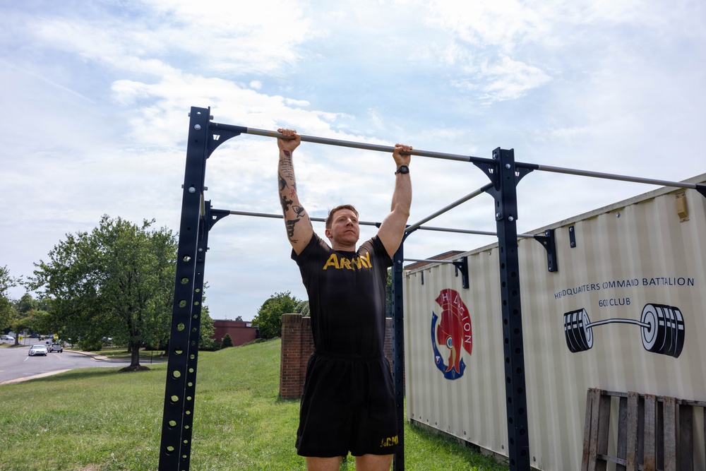 A male soldier wearing U.S. Army physical training attire is performing pull-ups on an outdoor exercise rack. The setting appears to be a military installation, with a shipping container in the background marked 'Headquarters Command Battalion 60i Club'. The image is taken outdoors during daytime under a partly cloudy sky.