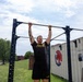 A male soldier wearing U.S. Army physical training attire is performing pull-ups on an outdoor exercise rack. The setting appears to be a military installation, with a shipping container in the background marked 'Headquarters Command Battalion 60i Club'. The image is taken outdoors during daytime under a partly cloudy sky.