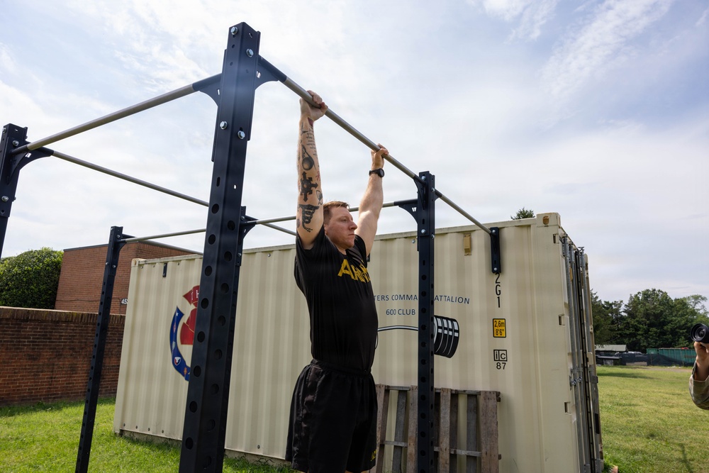 A tattooed individual wearing an Army-branded black shirt and shorts is performing a dead hang or pull-up on an outdoor metal pull-up rack. The setting appears to be a military installation, evidenced by a shipping container in the background labeled with battalion markings and an Army insignia. A photographer is partially visible on the right edge of the frame.