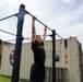 A tattooed individual wearing an Army-branded black shirt and shorts is performing a dead hang or pull-up on an outdoor metal pull-up rack. The setting appears to be a military installation, evidenced by a shipping container in the background labeled with battalion markings and an Army insignia. A photographer is partially visible on the right edge of the frame.