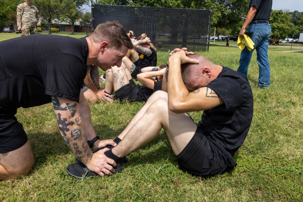 Military personnel in black PT (physical training) uniforms are performing sit-ups on a grass field, with one individual holding down another's feet to assist during the exercise. A tattooed instructor or partner kneels to anchor the feet of a shirtless soldier performing sit-ups. Additional personnel in the background suggest a group physical fitness assessment or training event.