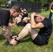 Military personnel in black PT (physical training) uniforms are performing sit-ups on a grass field, with one individual holding down another's feet to assist during the exercise. A tattooed instructor or partner kneels to anchor the feet of a shirtless soldier performing sit-ups. Additional personnel in the background suggest a group physical fitness assessment or training event.