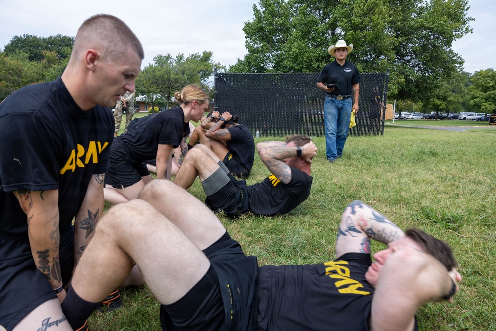 A group of U.S. Army soldiers in black PT (physical training) uniforms are performing sit-ups and partner-assisted exercises on a grass field. A civilian observer wearing a cowboy hat, black polo shirt, and jeans stands in the background holding a tablet or clipboard, apparently overseeing or evaluating the activity. The setting appears to be a military installation based on the fencing, buildings, and parked vehicles visible in the background.