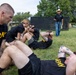 A group of U.S. Army soldiers in black PT (physical training) uniforms are performing sit-ups and partner-assisted exercises on a grass field. A civilian observer wearing a cowboy hat, black polo shirt, and jeans stands in the background holding a tablet or clipboard, apparently overseeing or evaluating the activity. The setting appears to be a military installation based on the fencing, buildings, and parked vehicles visible in the background.
