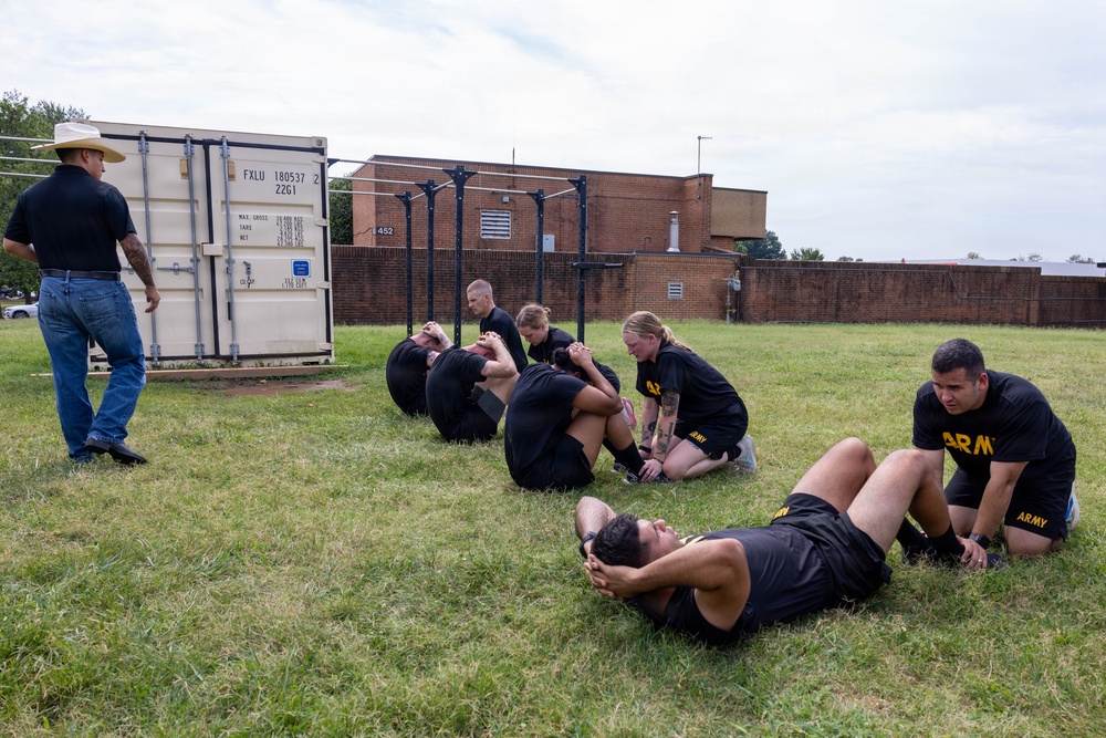 A group of U.S. Army soldiers in black PT (physical training) uniforms are performing sit-ups on a grass field while partners hold their feet. A civilian instructor wearing a cowboy hat, jeans, and boots observes the exercise from the left. The setting appears to be a military installation with a shipping container, pull-up bars, and brick buildings visible in the background.