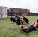 A group of U.S. Army soldiers in black PT (physical training) uniforms are performing sit-ups on a grass field while partners hold their feet. A civilian instructor wearing a cowboy hat, jeans, and boots observes the exercise from the left. The setting appears to be a military installation with a shipping container, pull-up bars, and brick buildings visible in the background.