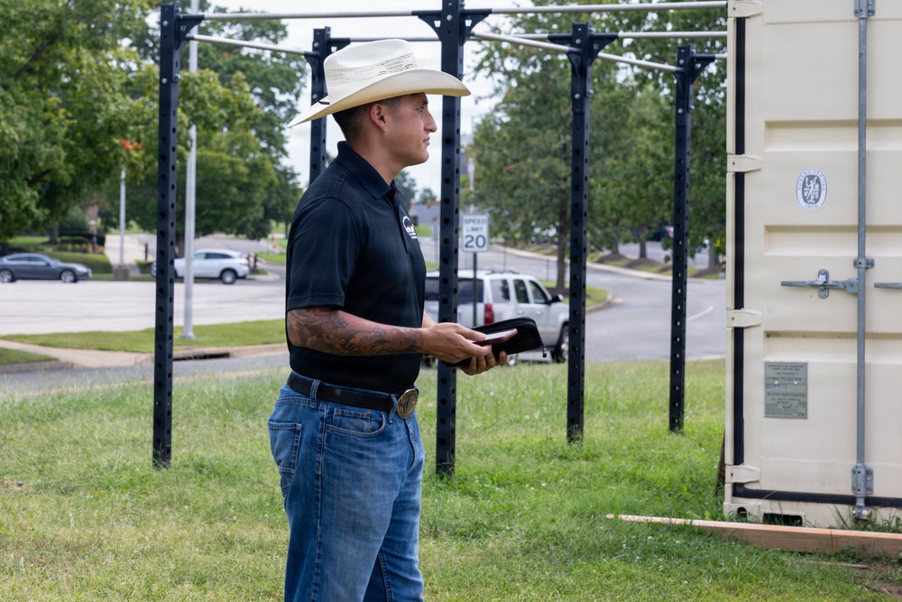 A man wearing a white cowboy hat, black polo shirt, and jeans stands on a grass area outdoors, holding what appears to be a wallet or small book. He has visible tattoos on his forearm and is wearing a decorative belt buckle. Behind him are outdoor pull-up/exercise racks and a beige shipping container, with a road and parked vehicles visible in the background.