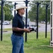 A man wearing a white cowboy hat, black polo shirt, and jeans stands on a grass area outdoors, holding what appears to be a wallet or small book. He has visible tattoos on his forearm and is wearing a decorative belt buckle. Behind him are outdoor pull-up/exercise racks and a beige shipping container, with a road and parked vehicles visible in the background.