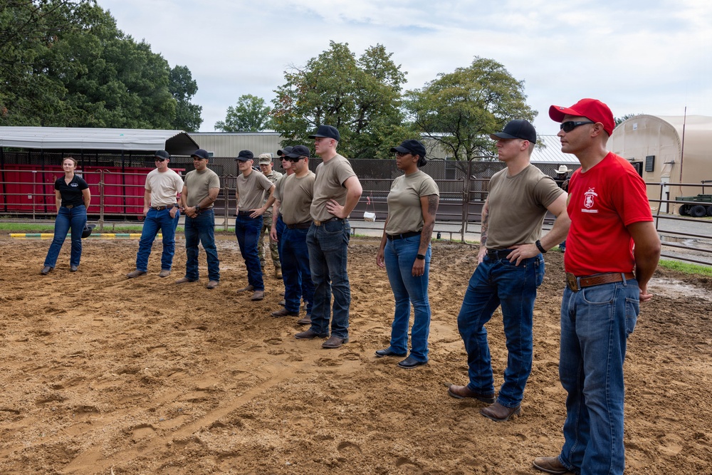 A group of approximately ten individuals, most wearing tan military-style t-shirts and jeans with dark baseball caps, are standing in a line on a sandy/dirt surface that appears to be a training or equestrian arena. They appear to be receiving instruction or a briefing, with one person in a red shirt and red cap standing at the front-right, likely in an instructor or leadership role. The setting includes fenced enclosures, a red barn structure, and trees in the background.