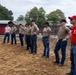 A group of approximately ten individuals, most wearing tan military-style t-shirts and jeans with dark baseball caps, are standing in a line on a sandy/dirt surface that appears to be a training or equestrian arena. They appear to be receiving instruction or a briefing, with one person in a red shirt and red cap standing at the front-right, likely in an instructor or leadership role. The setting includes fenced enclosures, a red barn structure, and trees in the background.