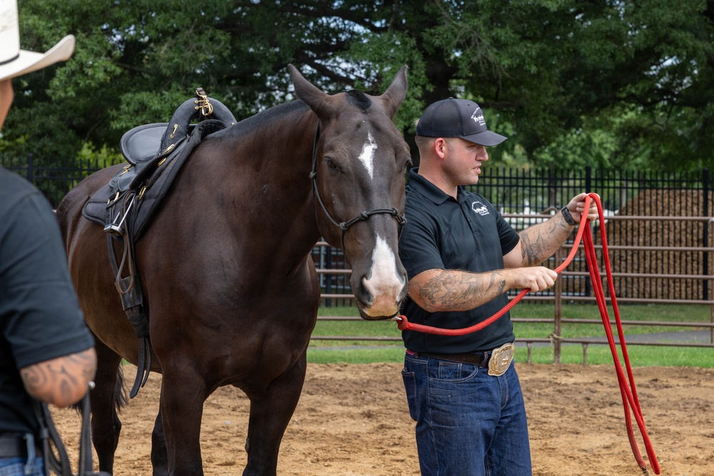 A tattooed man wearing a dark polo shirt, jeans, and a baseball cap is handling a dark brown saddled horse in an outdoor dirt arena. He holds a red lead rope while standing beside the horse, which has a white blaze marking on its face. A second person in a white cowboy hat is partially visible on the left edge of the frame.