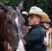 A young man wearing a white cowboy hat and black polo shirt stands beside a dark brown horse with a white blaze marking, fitted with a red lead rope. Behind him, a smiling young woman is adjusting or fastening a riding helmet. The setting appears to be a horse stable or equestrian facility with wooden fencing and trees visible in the background.