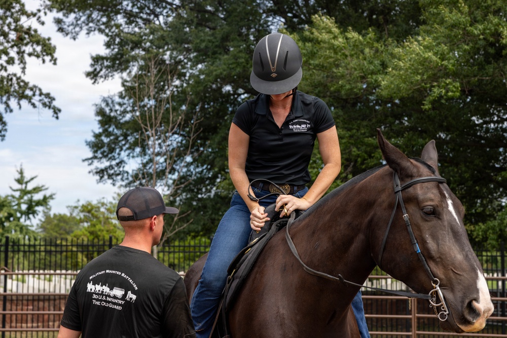 A woman wearing an equestrian helmet and black polo shirt is seated on a dark brown horse inside a fenced riding arena. A man wearing a black 'Military Mounted Battalion, 3d U.S. Infantry, The Old Guard' t-shirt stands beside her, appearing to provide instruction or assistance. The setting appears to be an outdoor equestrian facility with trees and a black iron fence visible in the background.