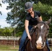 A woman wearing a black equestrian helmet and black polo shirt is riding a dark brown horse with a white blaze marking on its face. She is leaning forward and appears to be patting or stroking the horse's neck in an enclosed riding area. The setting appears to be an outdoor equestrian facility with metal fencing and mature trees in the background.