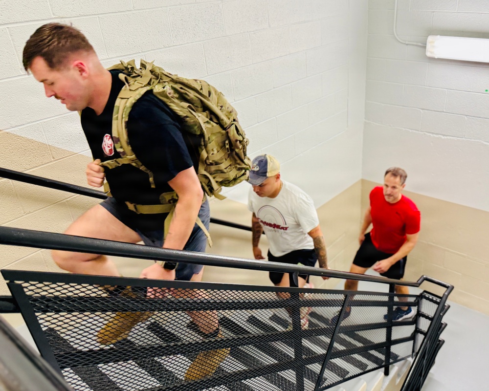 Three men are climbing an indoor metal staircase in what appears to be a fitness or training exercise. The lead man is wearing a tactical/military-style weighted rucksack over a black t-shirt, suggesting a rucking workout. The other two men follow behind in casual athletic wear.