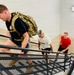 Three men are climbing an indoor metal staircase in what appears to be a fitness or training exercise. The lead man is wearing a tactical/military-style weighted rucksack over a black t-shirt, suggesting a rucking workout. The other two men follow behind in casual athletic wear.