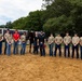 A group of approximately 14 people pose for a group photo in a sandy equestrian arena or paddock, with a dark horse held by two individuals in black shirts at the center. The participants appear to be military personnel in tan/khaki attire alongside civilian instructors, suggesting a equine therapy or horsemanship training program. A trailer in the background bears partial text reading '3d U.S. Inf...' indicating a U.S. Army unit affiliation.