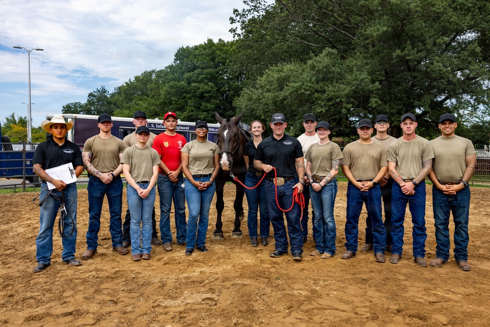 A group of approximately 14 individuals, appearing to be military personnel in tan/olive uniforms and civilians in black polo shirts, pose for a group photo with a dark horse on a sandy arena. A trailer in the background bears text referencing the 'U.S. Infantry Regiment.' The setting appears to be a military equestrian facility or training area.