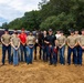 A group of approximately 14 individuals, appearing to be military personnel in tan/olive uniforms and civilians in black polo shirts, pose for a group photo with a dark horse on a sandy arena. A trailer in the background bears text referencing the 'U.S. Infantry Regiment.' The setting appears to be a military equestrian facility or training area.