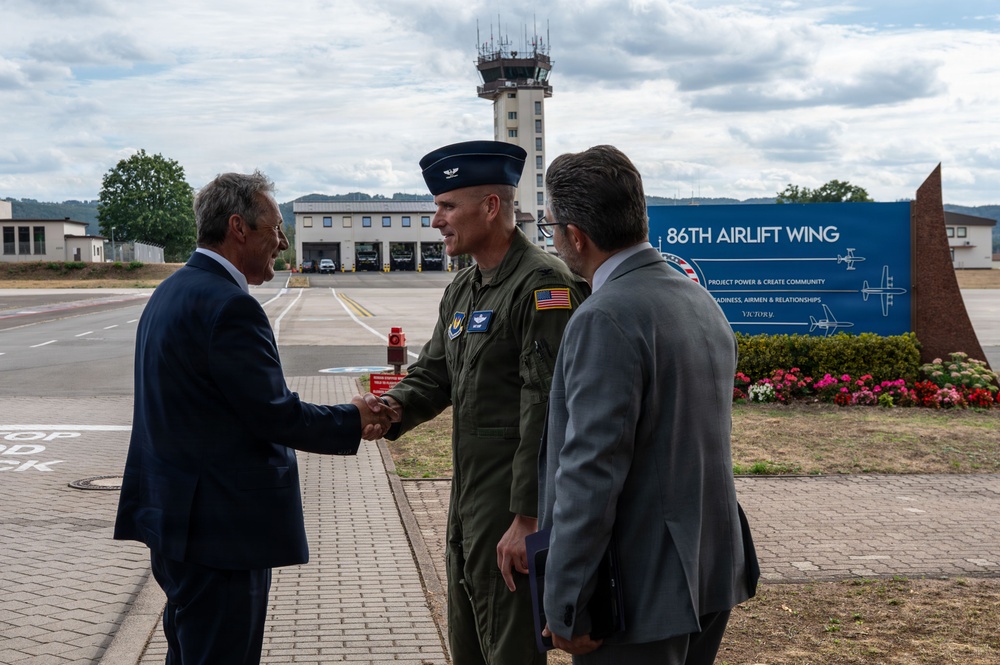 A U.S. Air Force officer in olive green flight suit is shaking hands with a civilian in a dark suit at the 86th Airlift Wing, likely at Ramstein Air Base in Germany. A third individual in a gray suit stands nearby observing the greeting. The setting appears to be an official arrival or welcoming ceremony on the flight line.