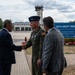A U.S. Air Force officer in olive green flight suit is shaking hands with a civilian in a dark suit at the 86th Airlift Wing, likely at Ramstein Air Base in Germany. A third individual in a gray suit stands nearby observing the greeting. The setting appears to be an official arrival or welcoming ceremony on the flight line.