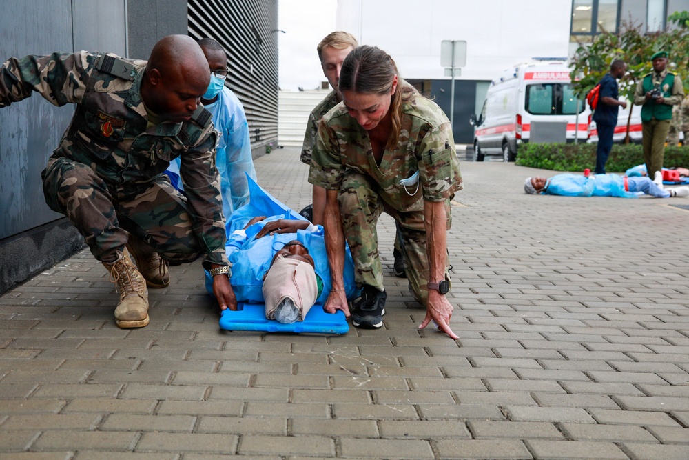 The image shows what appears to be a mass casualty exercise or training drill involving military and medical personnel. Multiple responders in camouflage uniforms are attending to a simulated casualty lying on a blue mat on a paved surface. An ambulance and additional personnel are visible in the background, along with other simulated casualties on the ground.