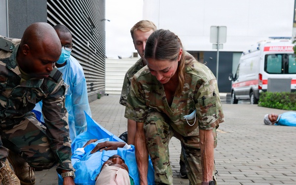 The image shows what appears to be a mass casualty exercise or training drill involving military and medical personnel. Multiple responders in camouflage uniforms are attending to a simulated casualty lying on a blue mat on a paved surface. An ambulance and additional personnel are visible in the background, along with other simulated casualties on the ground.
