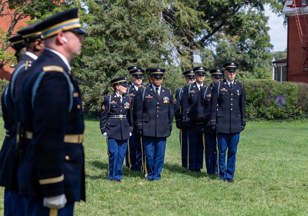 A formal U.S. Army military ceremony is taking place on a grass field, with soldiers in dress blue uniforms standing at attention in formation. A senior officer in the foreground appears to be inspecting or overseeing the assembled troops. The setting includes mature trees and a red brick building in the background, suggesting a military installation or historic post.