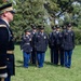 A formal U.S. Army military ceremony is taking place on a grass field, with soldiers in dress blue uniforms standing at attention in formation. A senior officer in the foreground appears to be inspecting or overseeing the assembled troops. The setting includes mature trees and a red brick building in the background, suggesting a military installation or historic post.
