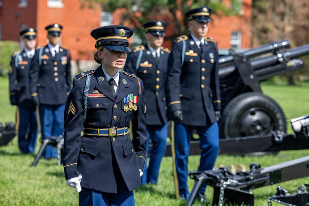 A group of U.S. Army soldiers in dress blue uniforms are participating in a formal military ceremony, likely a cannon salute or honor guard event. A female sergeant leads the formation in the foreground, wearing medals and a distinctive blue cord (aiguillette). The setting appears to be a military installation with red brick buildings visible in the background.