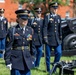 A group of U.S. Army soldiers in dress blue uniforms are participating in a formal military ceremony, likely a cannon salute or honor guard event. A female sergeant leads the formation in the foreground, wearing medals and a distinctive blue cord (aiguillette). The setting appears to be a military installation with red brick buildings visible in the background.
