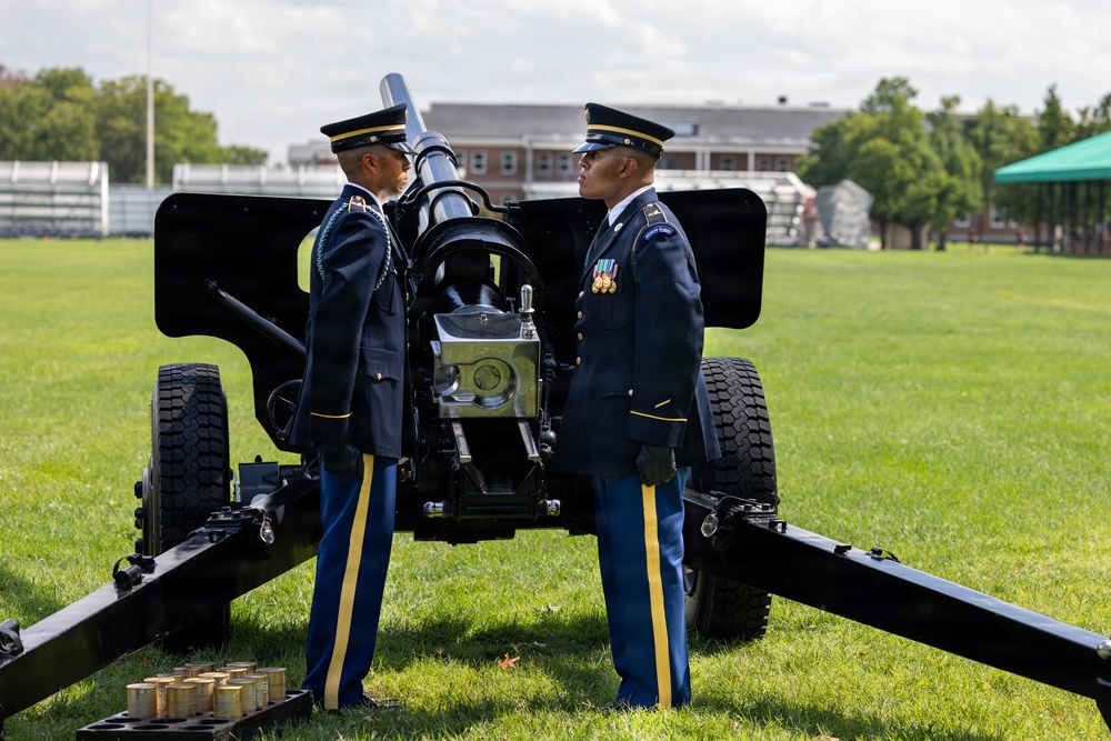 Two U.S. Army soldiers in dress blue uniforms stand at attention on either side of a ceremonial artillery cannon on a green parade field. The setting appears to be a formal military ceremony, likely a cannon salute or honors detail, at what appears to be a U.S. Army installation. Several artillery rounds are visible on the ground near the cannon's trail.