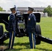 Two U.S. Army soldiers in dress blue uniforms stand at attention on either side of a ceremonial artillery cannon on a green parade field. The setting appears to be a formal military ceremony, likely a cannon salute or honors detail, at what appears to be a U.S. Army installation. Several artillery rounds are visible on the ground near the cannon's trail.