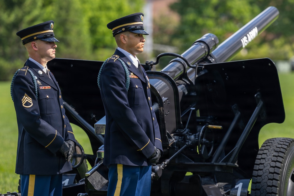Two U.S. Army soldiers in dress blue uniforms stand at attention beside a ceremonial artillery cannon marked 'US ARMY'. The setting appears to be a formal military ceremony, likely a salute battery or honors detail, conducted outdoors on a grassy area. Both soldiers are wearing ceremonial dress uniforms with distinctive aiguillettes and service decorations.