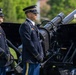 Two U.S. Army soldiers in dress blue uniforms stand at attention beside a ceremonial artillery cannon marked 'US ARMY'. The setting appears to be a formal military ceremony, likely a salute battery or honors detail, conducted outdoors on a grassy area. Both soldiers are wearing ceremonial dress uniforms with distinctive aiguillettes and service decorations.