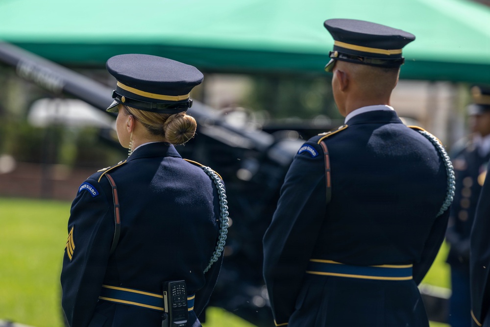 Two U.S. Army Honor Guard soldiers in dress blue uniforms are standing at attention during a formal military ceremony. Both wear distinctive Honor Guard shoulder patches and ceremonial aiguillettes. Artillery pieces and a green canopy tent are visible in the background.
