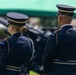 Two U.S. Army Honor Guard soldiers in dress blue uniforms are standing at attention during a formal military ceremony. Both wear distinctive Honor Guard shoulder patches and ceremonial aiguillettes. Artillery pieces and a green canopy tent are visible in the background.