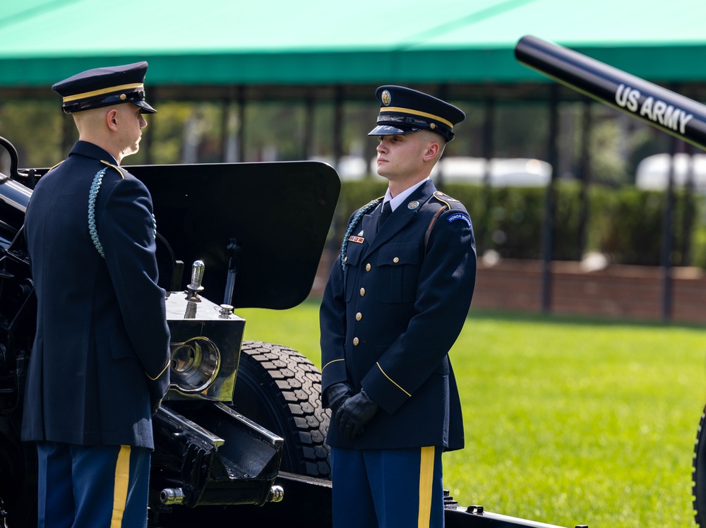 Two U.S. Army soldiers in dress blue uniforms stand at attention beside a ceremonial artillery cannon during what appears to be a formal military ceremony. The soldier on the right displays an 'Honor Guard' shoulder tab and aiguillette, indicating this is likely a salute battery or honor guard detail. The setting appears to be an outdoor ceremonial ground with a green canopy tent visible in the background.