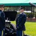 Two U.S. Army soldiers in dress blue uniforms stand at attention beside a ceremonial artillery cannon during what appears to be a formal military ceremony. The soldier on the right displays an 'Honor Guard' shoulder tab and aiguillette, indicating this is likely a salute battery or honor guard detail. The setting appears to be an outdoor ceremonial ground with a green canopy tent visible in the background.