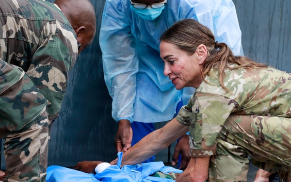 Military and medical personnel are attending to a patient lying on a blue medical drape on the ground. At least four individuals are visible, including personnel in camouflage uniforms and one person in blue surgical gown and mask. The scene appears to depict a field medical training exercise or emergency casualty care situation.