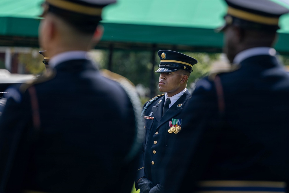 The image shows U.S. Army soldiers in dress blue uniforms during what appears to be a formal military ceremony or parade. A central soldier in sharp focus stands at attention wearing medals, ribbons, and a service cap with gold insignia. Two other soldiers in the foreground are blurred, creating a depth-of-field effect that draws attention to the central figure.