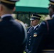 The image shows U.S. Army soldiers in dress blue uniforms during what appears to be a formal military ceremony or parade. A central soldier in sharp focus stands at attention wearing medals, ribbons, and a service cap with gold insignia. Two other soldiers in the foreground are blurred, creating a depth-of-field effect that draws attention to the central figure.