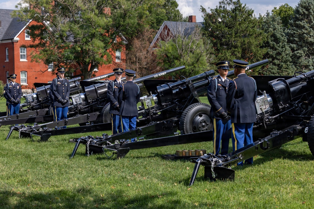 A formal US Army ceremonial cannon salute is being conducted on a grassy field, with uniformed soldiers standing at attention beside multiple black artillery pieces. The soldiers are dressed in Army Service Uniforms (blue dress uniforms) with gold striping on the trousers, indicating a formal military ceremony. Red brick buildings and mature trees are visible in the background, suggesting a military installation or post.