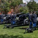 A formal US Army ceremonial cannon salute is being conducted on a grassy field, with uniformed soldiers standing at attention beside multiple black artillery pieces. The soldiers are dressed in Army Service Uniforms (blue dress uniforms) with gold striping on the trousers, indicating a formal military ceremony. Red brick buildings and mature trees are visible in the background, suggesting a military installation or post.
