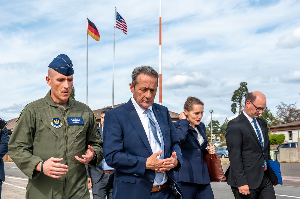 A group of individuals, including a U.S. Air Force officer in flight suit and several civilians in business attire, are walking together outdoors at what appears to be a U.S. military installation in Germany. Both a German flag and a U.S. flag are visible flying on flagpoles in the background. The scene suggests an official visit or delegation walkthrough at a joint military facility.