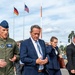 A group of individuals, including a U.S. Air Force officer in flight suit and several civilians in business attire, are walking together outdoors at what appears to be a U.S. military installation in Germany. Both a German flag and a U.S. flag are visible flying on flagpoles in the background. The scene suggests an official visit or delegation walkthrough at a joint military facility.
