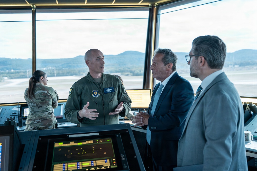 The image shows what appears to be a military air traffic control tower briefing, where a U.S. Air Force officer in a flight suit is speaking with two civilian officials in business attire. A female soldier in camouflage uniform is visible in the background working at the control station. The setting features large panoramic windows overlooking an airfield with mountains visible in the distance.
